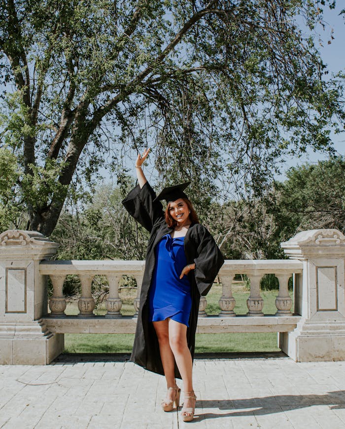 Full body of cheerful female student in black square academic cap and cloak raising arm and looking at camera with bright smile while standing near stone fence under green tree